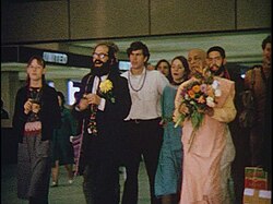 Allen Ginsberg ร่วมกับสวามีในชุดสีส้มและกลุ่มผู้ติดตามที่สนามบิน